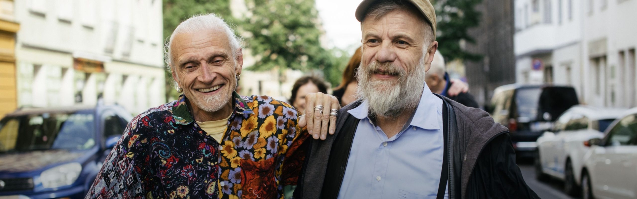 Two older adult men smiling arm in arm on a street